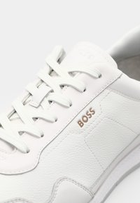 Baskets blanches en cuir texturé, avec des lacets plats blancs, une semelle en caoutchouc, et un logo "BOSS" marron discret.