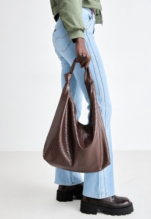 Person, der hellblaue Jeans und dunkelbraune Stiefel trägt, hält eine große, braune, gewebte Leder-Umhängetasche am Griff.