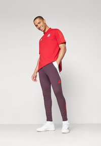 Rød polo skjorte med logo og tre striber, parret med mørkelilla tapered bukser med hvide og røde accenter og hvide sportssko.