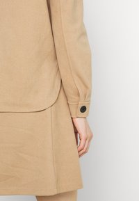 Il capo beige presenta una texture liscia, maniche aderenti con un bottone nero e una gonna a trapezio con orlo arrotondato.