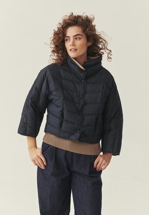 Žena s kučeravými vlasmi oblečená v krátkej čiernej vatovanej bunde, pod ňou béžový sveter a tmavé široké nohavice, stojaca so založenými rukami na bokoch.