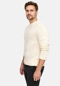 Maglione lavorato a maglia color crema con un motivo a rombi, scollo a girocollo e polsini a costine. Abbinato a pantaloni scuri, il modello sta in posa di profilo.