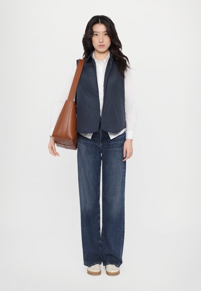 Marineblauwe mouwloze vest over een witte blouse, gecombineerd met wijde blauwe jeans en lichtgekleurde schoenen, terwijl een bruine lederen tas wordt vastgehouden.