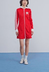 Rode sportieve jurk met een witte rits aan de voorkant, contrasterende witte kraag en manchetten, en drie witte strepen op elke mouw. Adidas-logo op de borst.