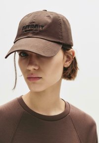 Brown cotton baseball cap with embroidered "DETROIT" text, featuring a curved brim and adjustable back strap. Model wears a matching brown top.