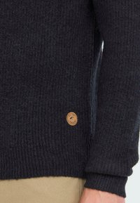 Gebreide donkergrijze trui met ribbeltextuur, voorzien van een klein tan leren logo-oproepje aan de linkerkant bij de zoom.