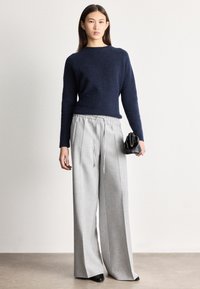 Sieviete stāv, valkājot tumši zilas krāsas džemperi, gaiši pelēkas krāsas platu kāju bikses ar auklu, melnus kurpes, turot mazā melnā somiņu pret vienkāršu fona.