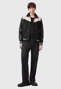 Zwarte en crème varsityjack met een kraag, drukknoopsluiting en gestreepte manchetten; gedragen over een zwarte ribbed shirt en gecombineerd met zwarte broek.