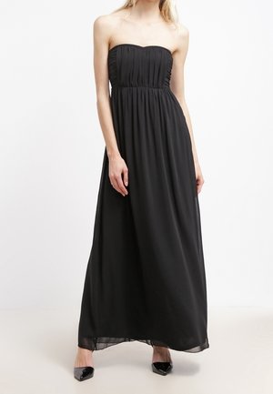 Femme portant une robe maxi noire sans bretelles avec un corsage plissé et des escarpins noirs pointus, debout sur un fond blanc.
