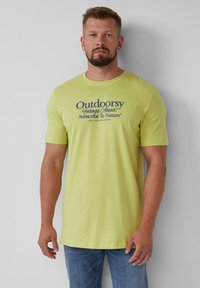 Geel katoenen T-shirt met marineblauwe grafische tekst op de voorkant. Korte mouwen en een relaxte pasvorm, gecombineerd met lichtblauwe jeans.