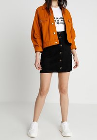 Chaqueta de mezclilla naranja, camisa blanca con texto, falda de ante negra con botones y zapatillas blancas. El conjunto presenta colores y texturas contrastantes.