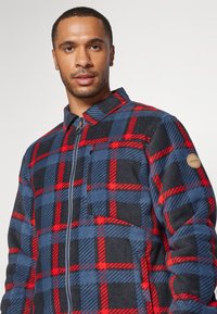 Fleecová bunda s modro-červeným a čiernym károvaným vzorom, so zipsom, špicatým golierom a označením na rukáve.