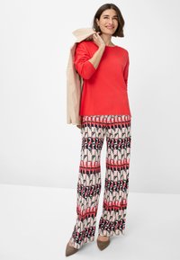 Top rosso a maniche lunghe abbinato a pantaloni larghi a fantasia nei toni del rosa, rosso e nero. La modella tiene una giacca beige. Tacchi marroni.