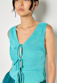 Mujer con blusa sin mangas de lino turquesa con tres lazos delanteros y pendientes de aro dorados con charms turquesas.