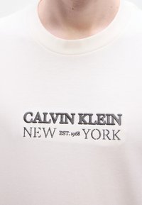 Βαμβακερό μπλουζάκι, εκρού χρώμα, λαιμόκοψη με ριμπ ύφανση, διαθέτει κεντημένο κείμενο "CALVIN KLEIN NEW YORK EST. 1968" σε γκρι και μαύρο.