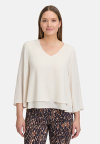 Blusa beige con scollatura a V e maniche in stile kimono; texture liscia; design a strati. Abbinata a pantaloni a motivi marrone scuro e crema.