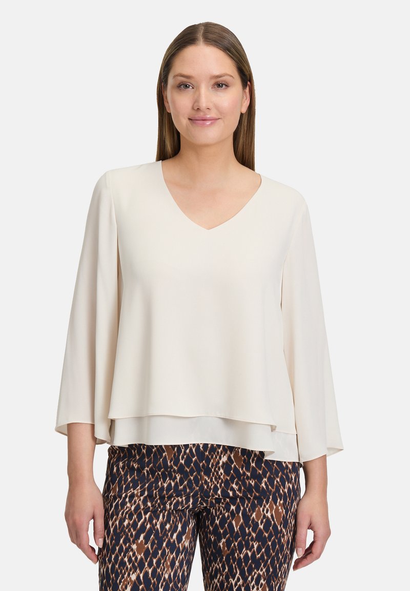 Blusa beige con scollatura a V e maniche in stile kimono; texture liscia; design a strati. Abbinata a pantaloni a motivi marrone scuro e crema.