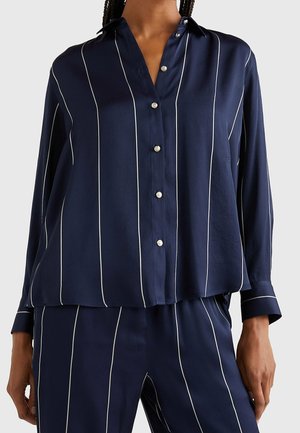 Woman wearing navy blue long-sleeve button-up shirt and matching pants with thin vertical white stripes, standing against a plain background.