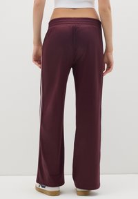 Person in burgunderfarbigen Hosen mit weißen Seitenstreifen, weißem Crop-Top und weißen Sneakers mit Gummisohlen, der vor einer schlichten hellen Wand steht.