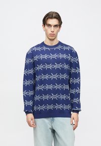 Πλεκτό μπλε μπλουζάκι με επαναλαμβανόμενο σχέδιο από σύρμα με αγκάθια σε ανοιχτό μπλε χρώμα. Ριμπ μανσέτες και τελείωμα, στρογγυλή λαιμόκοψη, κατασκευασμένο από μαλακό πλεκτό ύφασμα.