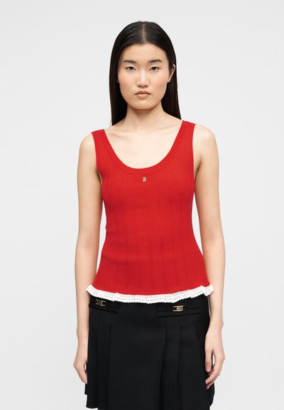 Γυναίκα με αμάνικη κόκκινη πλεκτή μπλούζα με λευκή δαντέλα και μαύρη πλισέ φούστα μπροστά σε γκρι φόντο.