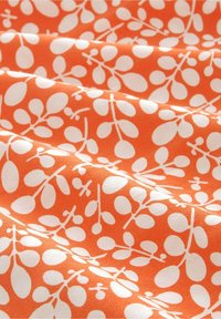 Tissu orange avec un motif de branche feuillue blanche, présenté avec des plis doux et une texture lisse.
