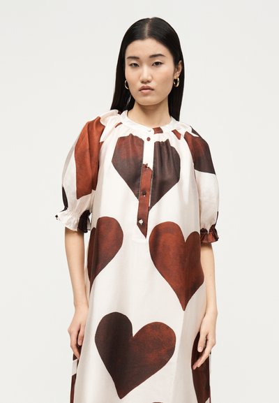 Νεαρή γυναίκα με μακριά σκούρα μαλλιά φοράει λευκό φόρεμα με μεγάλα σκούρα κόκκινα σχέδια καρδιάς και φουσκωτά μανίκια, στέκεται μπροστά σε απλό φωτεινό φόντο.