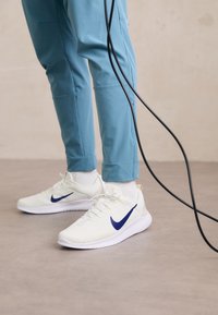 Zapatillas Nike blancas con un swoosh azul, que presentan una parte superior de malla texturizada y suela blanca, combinadas con pantalones ajustados de color azul claro.