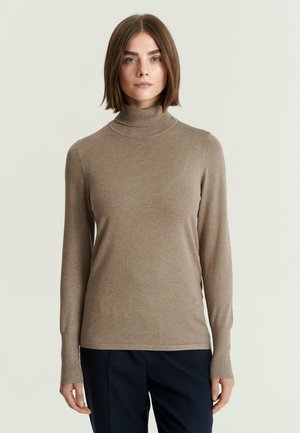 Femme aux cheveux bruns lisses portant un pull à col roulé beige et un pantalon sombre, debout devant un fond clair uni.
