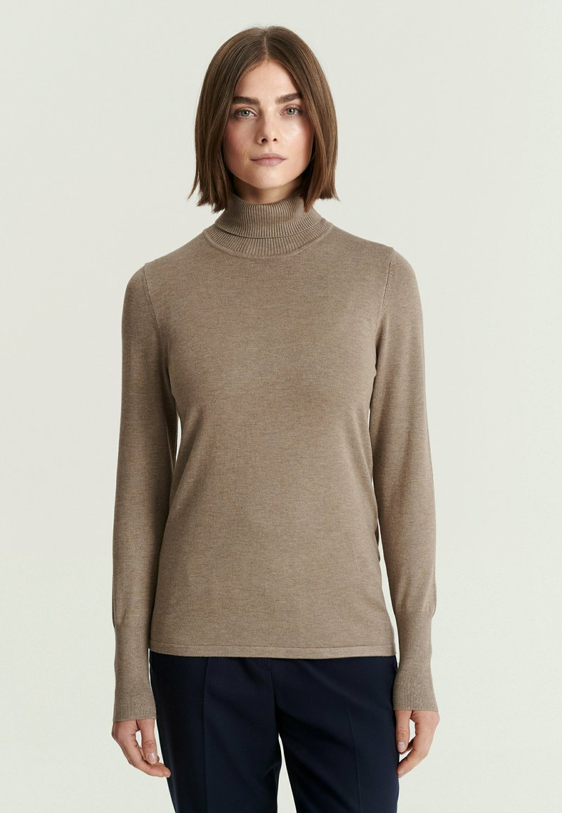 Femme aux cheveux bruns lisses portant un pull à col roulé beige et un pantalon sombre, debout devant un fond clair uni.