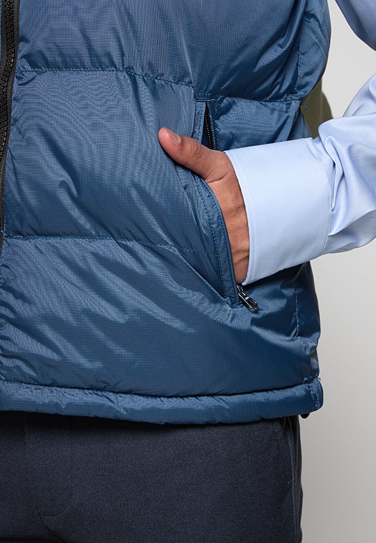 Gilet matelassé bleu marine présentant une finition texturée, une poche zippée sur le côté et un bas ourlé. Porté par-dessus une chemise à manches longues bleu clair.