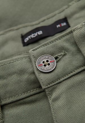 Tela de algodón verde oliva con un botón plateado, con texto "ombre wear" y acentos rojos. La etiqueta muestra tamaño H38. Vista en primer plano.