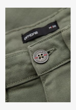 Tela de algodón verde oliva con un botón plateado, con texto "ombre wear" y acentos rojos. La etiqueta muestra tamaño H38. Vista en primer plano.