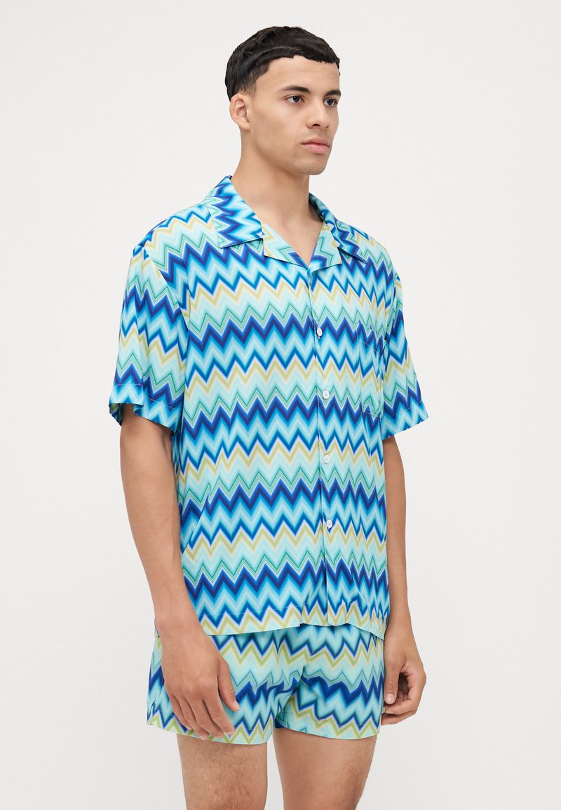 Pigiama a maniche corte con bottoni, in motivo zigzag con tonalità di blu e verde. Realizzato in tessuto leggero, include pantaloni shorts abbinati.