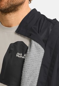 Schwarze Jacke mit grauem, flauschigem Innenfutter; verfügt über eine Reißverschlusstasche mit Logo, ein Netzschulterdetail und einen kontrastierenden Kragen.