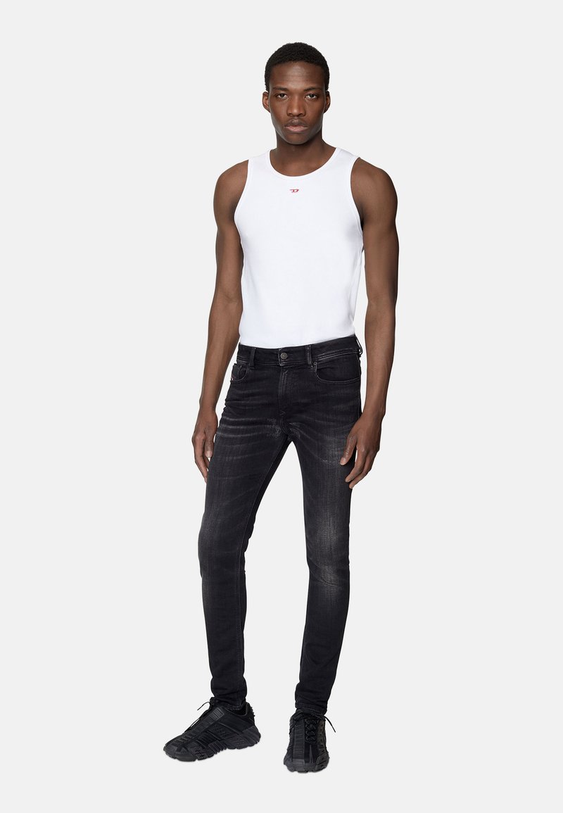 Jeune homme debout, portant un t-shirt sans manches blanc, un jean slim noir et des chaussures de sport noires, contre un fond clair uni.