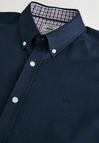 Marineblaue Knopf-auf-Shirt aus strapazierfähigem Stoff, mit einem karierter Muster im Kragen und einem kleinen Kronenemblem auf der Brust.