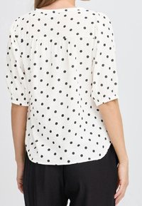 Femme portant un chemisier blanc à pois noirs avec des manches à coudes, associé à un pantalon noir, vue de dos.