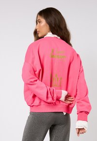 Ružová mikina s zeleným grafickým textom na chrbte, voľného strihu a s rebrovanými manžetami. Kombinovaná s trblietavými sivými legínami.