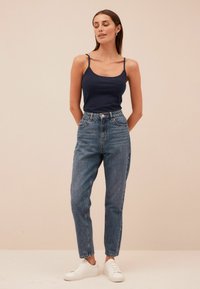 Top sin mangas azul marino, jeans de tiro alto de color azul claro, zapatillas blancas. La modelo está de pie con las manos en los bolsillos, fondo claro y neutro.