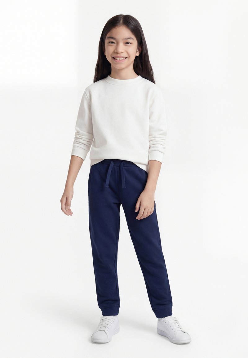 Fată zâmbitoare cu păr lung și închis la culoare, purtând un sweatshirt alb, pantaloni de trening bleumarin și adidași albi, stând în fața unui fundal alb.