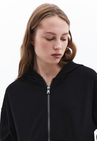 Felpa nera con zip, dalla texture morbida, grande cappuccio e zip argentata. Design semplice con vestibilità comoda e senza loghi visibili.