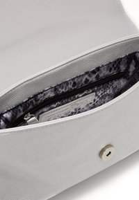 Stříbrná kožená taška uvnitř má šedou podšívku s hadím vzorem a zipovou kapsu. Obsahuje magnetické zapínání a hladký povrch.