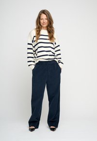 Gestreepte crème- en marineblauwe trui met geribbelde structuur, gecombineerd met marineblauwe wijduitlopende corduroy broek. Zwarte pumps met puntige neus. Effen achtergrond.