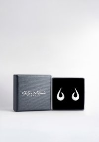 Söljtonade tårformade örhängen visade i en svart sammetspärla med en texturerad svart yta och vit logotyp.