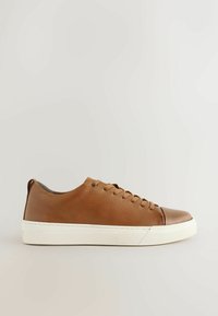 Zapatilla de cuero marrón con parte superior texturizada, puntera redondeada, suela de goma blanca y una puntera de cuero marrón en contraste. Presenta un diseño de cordones y una cinta de agarre en la parte trasera.