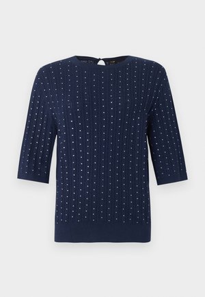 Kvinde stående med den ene hånd i lommen, iført en marineblå prikket sweater, hvide vidde bukser og mørkebrud sko.