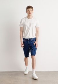 Hvit t-skjorte med korte ermer og en liten logo, blå denimshorts og hvite joggesko med synlige lisser og ankelstrømper. Enkelt hverdagsantrekk.