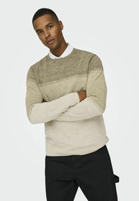 Beige gebreide trui met verschillende texturen en horizontale strepen; lange mouwen, ronde hals, gecombineerd met donkere broek.