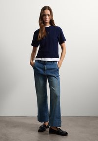 Pull en tricot côtelé bleu marine à manches courtes sur une chemise blanche, associé à un jean en denim bleu à jambes larges et des mocassins noirs à finition texturée.